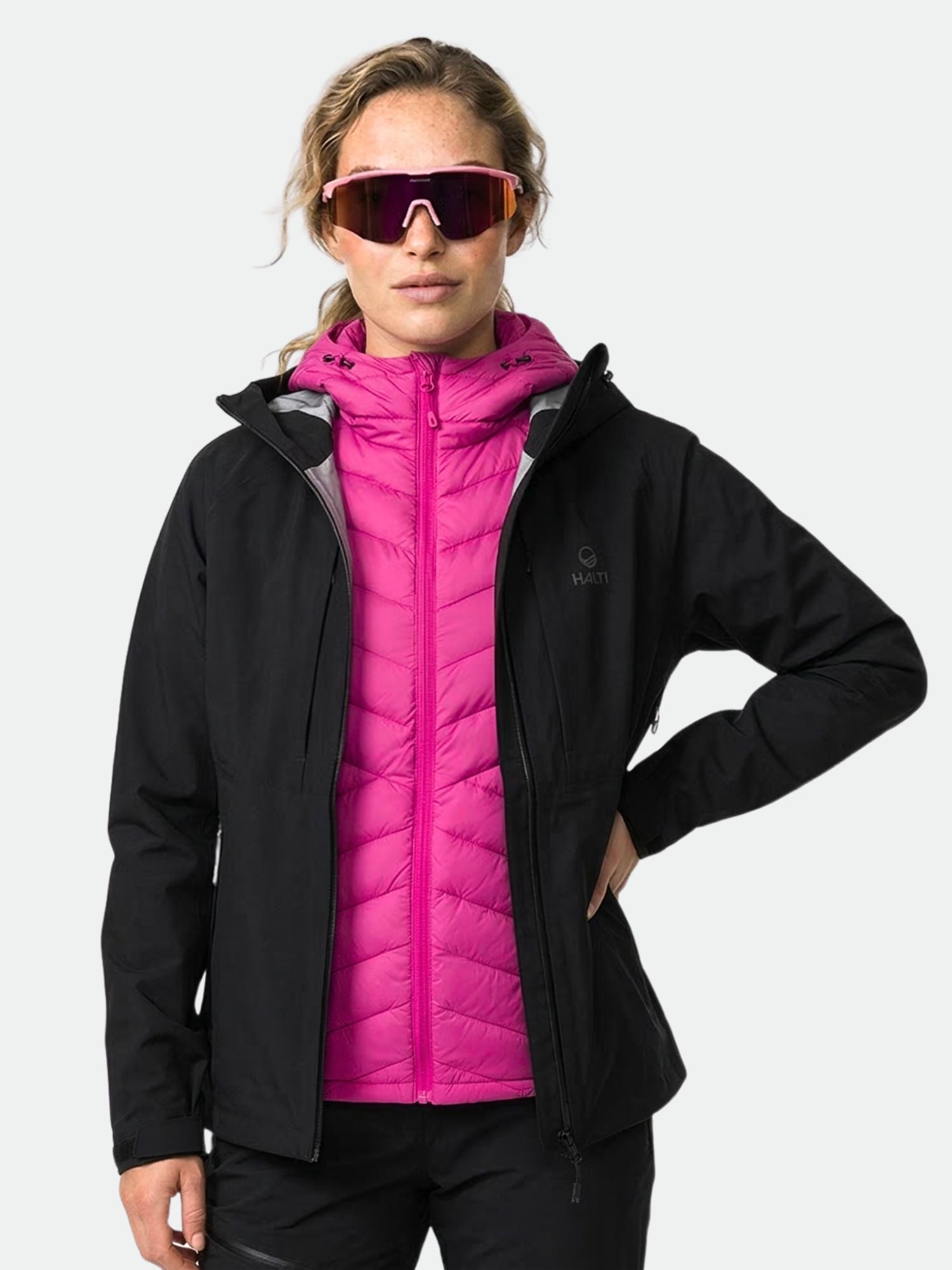 A woman wears the Halti Kaarna DX 3L Shell Jacket over a pink puffer and reflective sunglasses against a plain background.