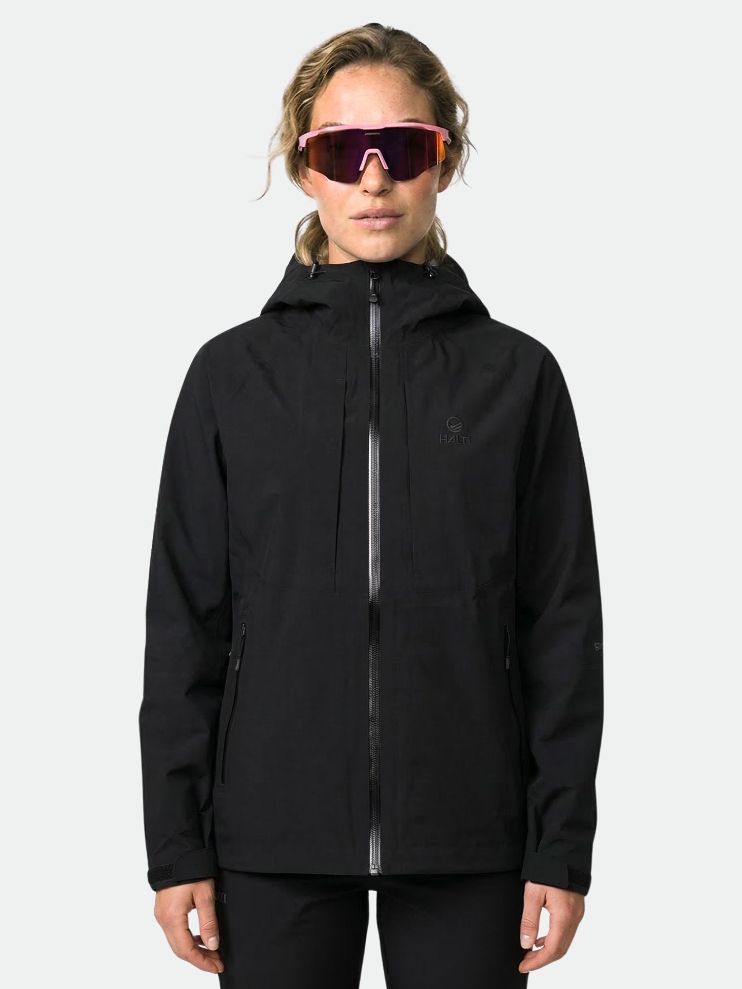 A person wears the Halti Kaarna DX 3L Shell Jacket Women's in black and large reflective sunglasses, facing forward.