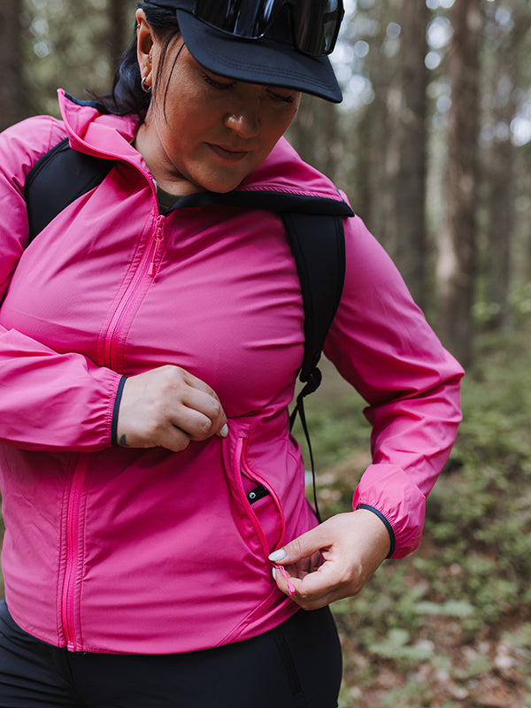 A woman in a pink Halti Pallas Plus Womens X-stretch Lite Jacket unzips a pocket outdoors in a wooded area.