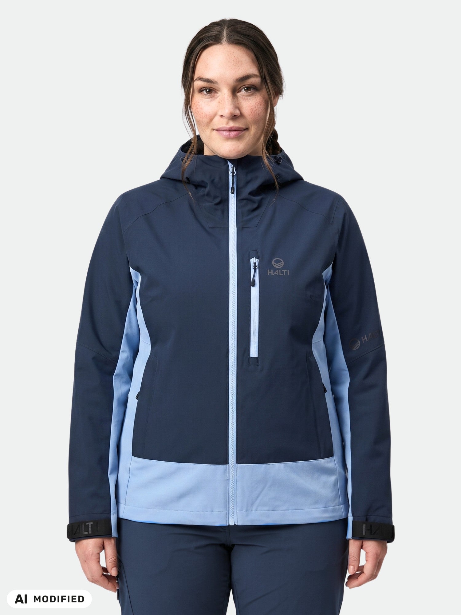 A woman wears the Halti Pallas Plus III Hybrid Jacket in blue with a hood, standing against a white background.