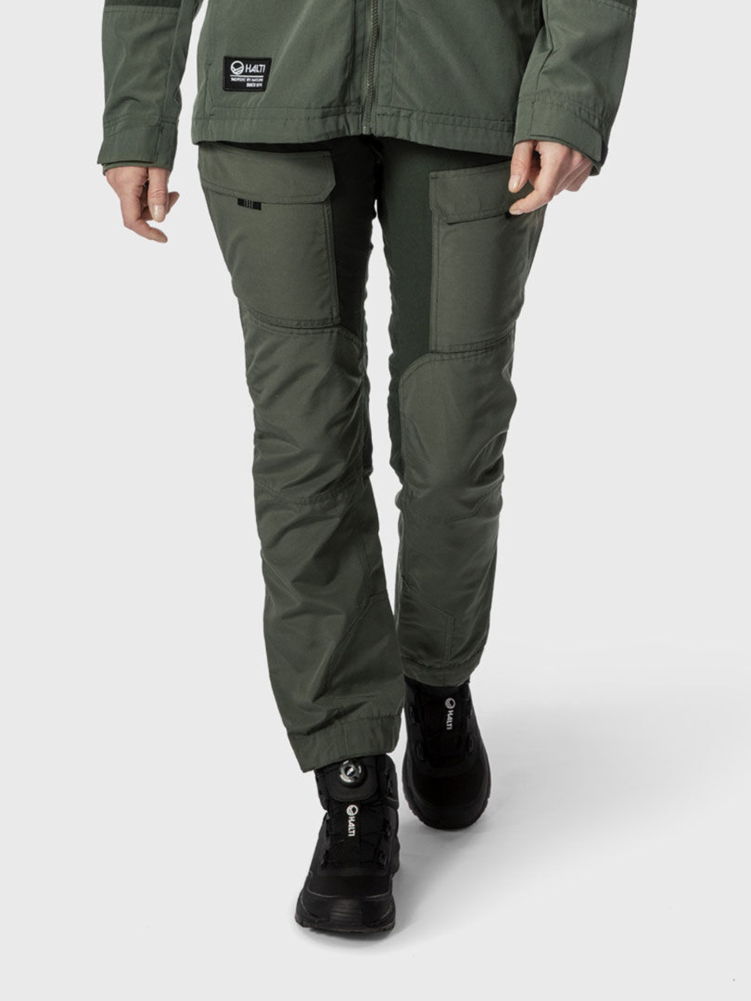 A woman in Halti Hiker Women's Ventilated Pants and a matching jacket, wearing black hiking shoes, walks on a plain background.