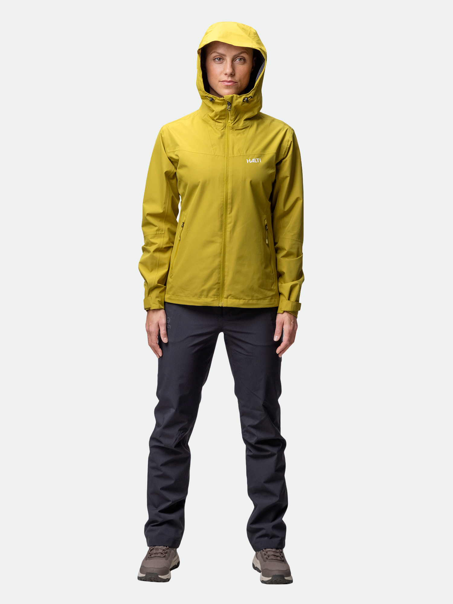 A woman wears the Halti Forter DX Shell Jacket in yellow with dark pants and brown shoes against a plain background.