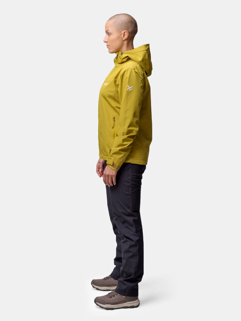 A woman stands in profile wearing a yellow Halti Forter DX Shell Jacket, black pants, and brown shoes against a plain background.