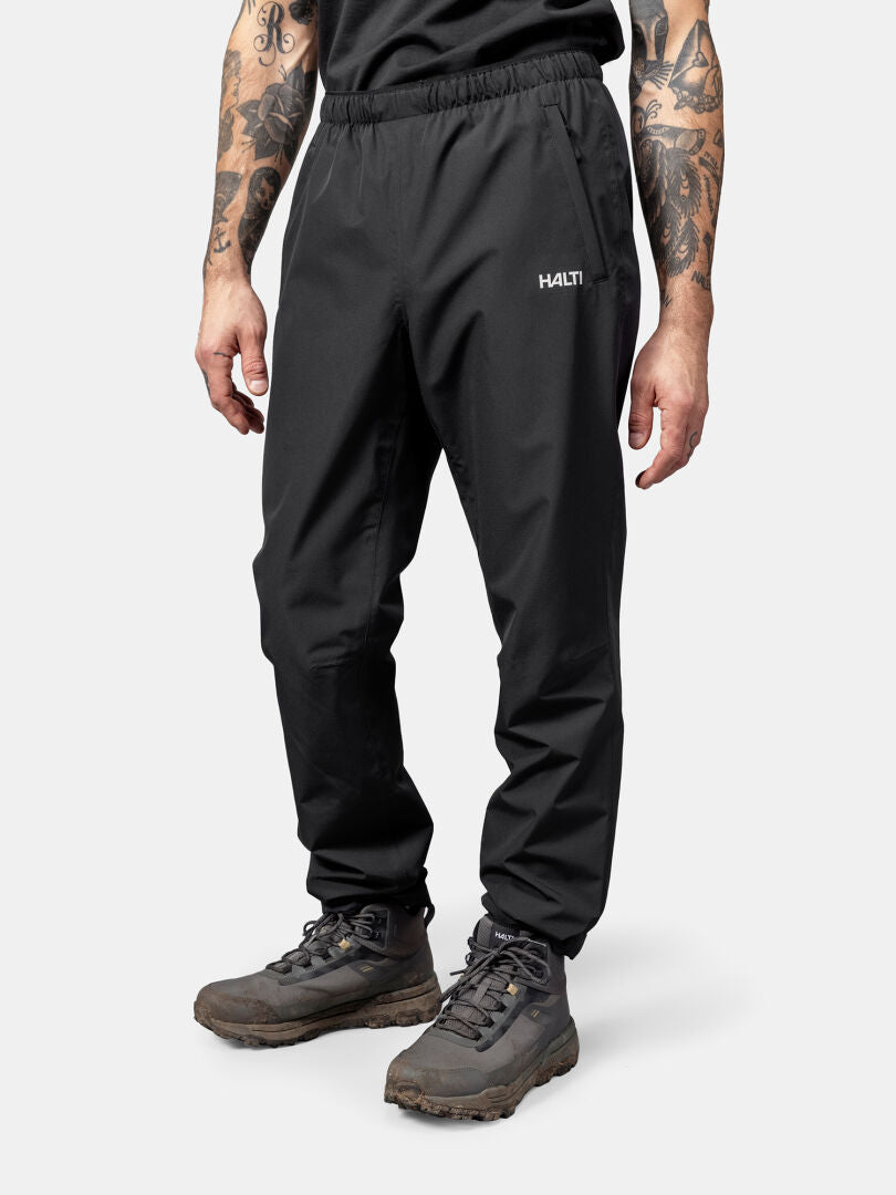 A person in Halti Forter DrymaxX Shell Pants, gray boots, and a black shirt stands against a plain background.