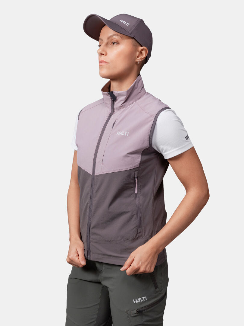 Person wearing Halti Pallas Evo X-stretch Vest for women, a white tee, green pants, and gray cap, hands near pockets.