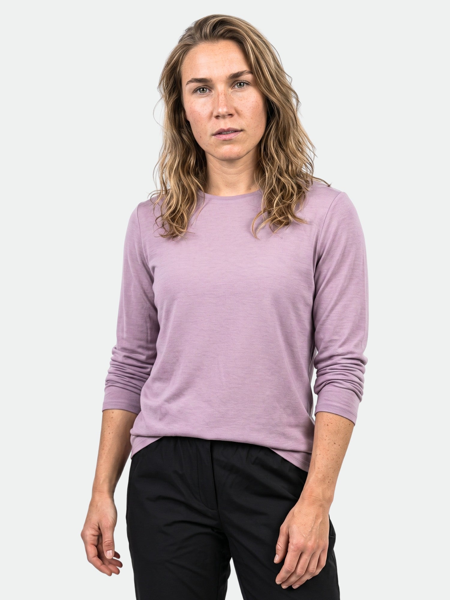Woman with wavy light brown hair wears Halti Tuntu II Long Sleeve Merino Shirt and black pants on a plain background.