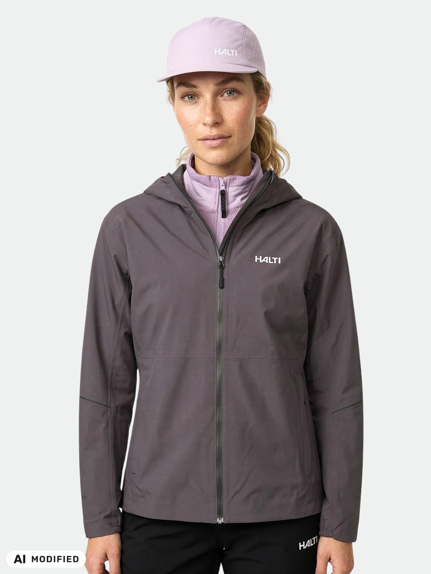 A woman wears the Halti Pisarat 2,5L DX Shell Jacket and a lilac cap, facing forward against a light gray background.