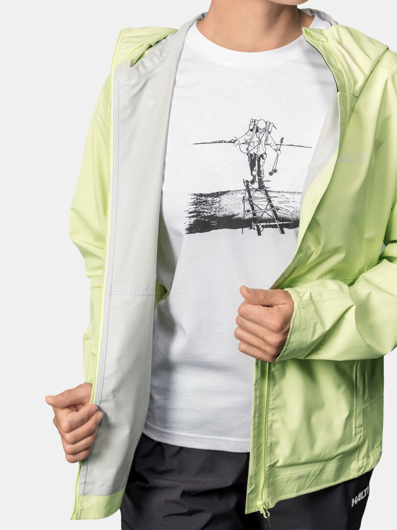 A person wears Halti's Pisarat 2.5L DX Shell Jacket Women's in light green over a white tee with a skier graphic.