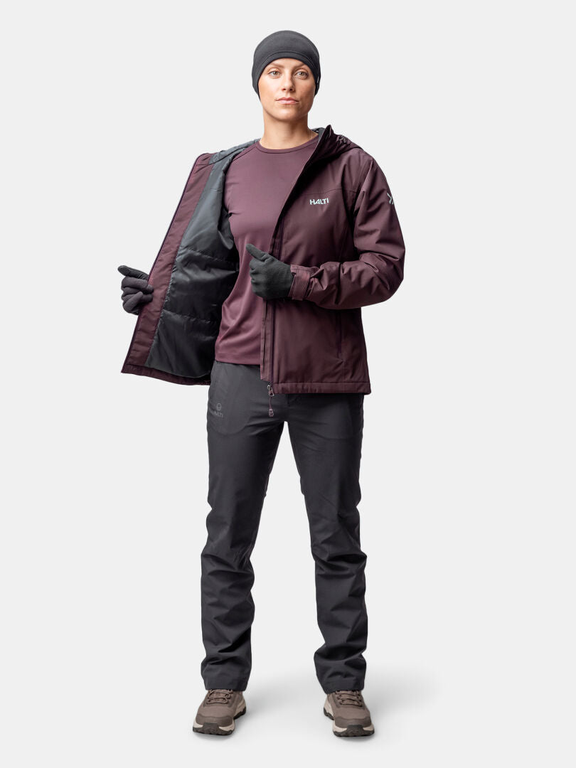 A woman in a maroon Halti Forter DX Warm Shell Jacket stands facing forward, wearing black pants, gloves, beanie, and hiking shoes.