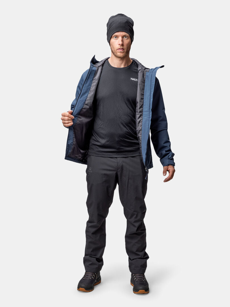 A man faces forward wearing the Halti Forter DX Warm Shell Jacket Men's, a beanie, black shirt, black pants, and hiking shoes.