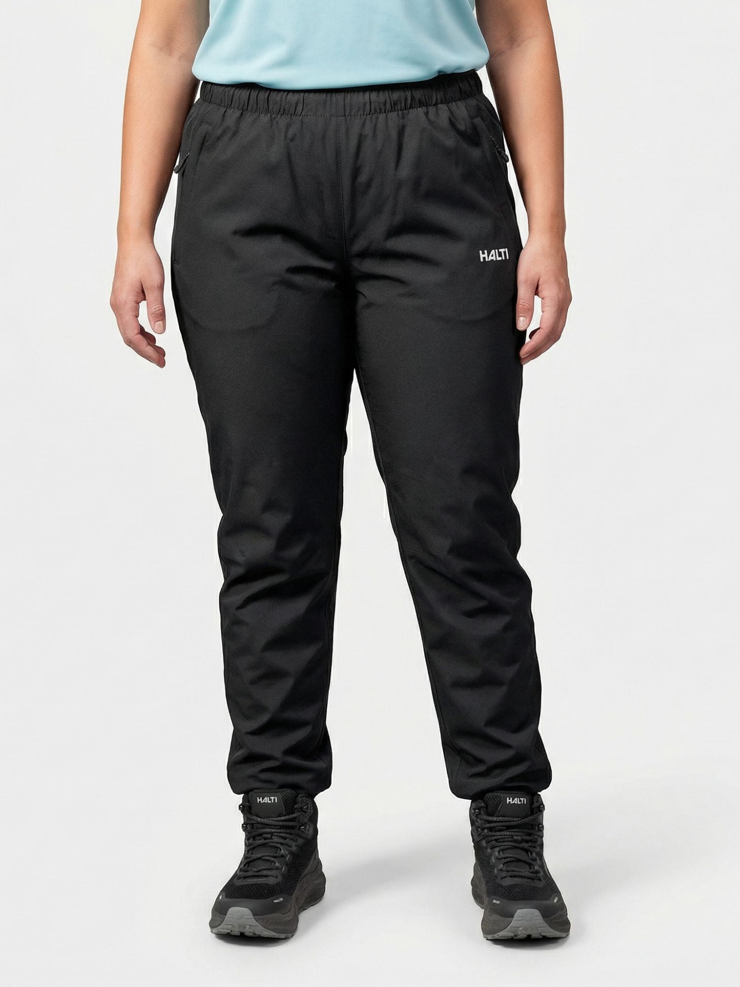 A person wears Halti Forter DX Shell Pants Women's Plus with black sneakers and a light blue shirt against a plain background.