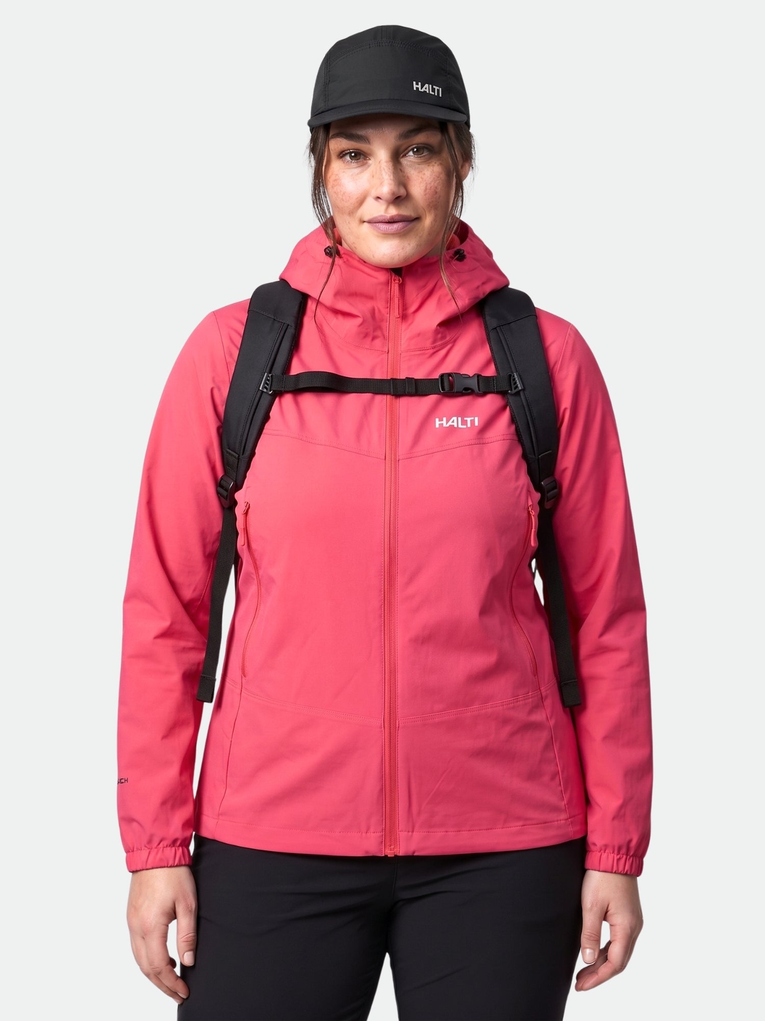 A woman in a red Halti Pallas Evo X-stretch Jacket Plus and black cap stands with a black backpack.