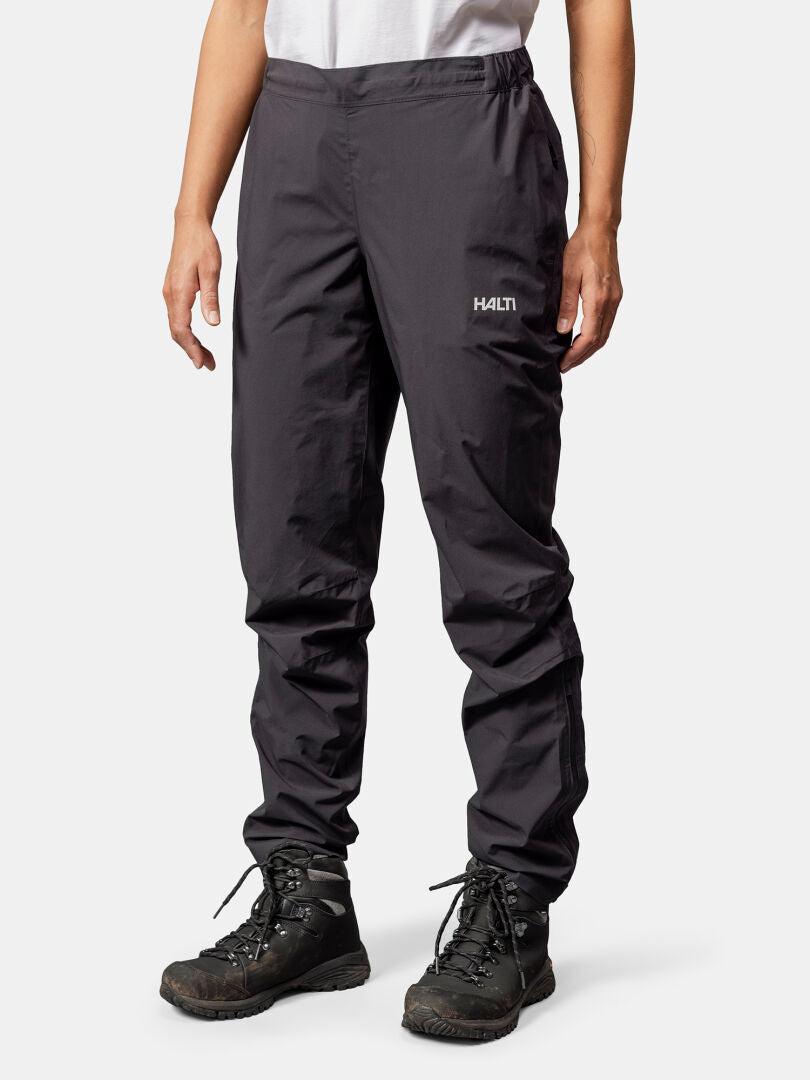 Person wearing Halti Pisarat 2,5L DX Shell Pants, black hiking boots, and a white shirt, against a white background.