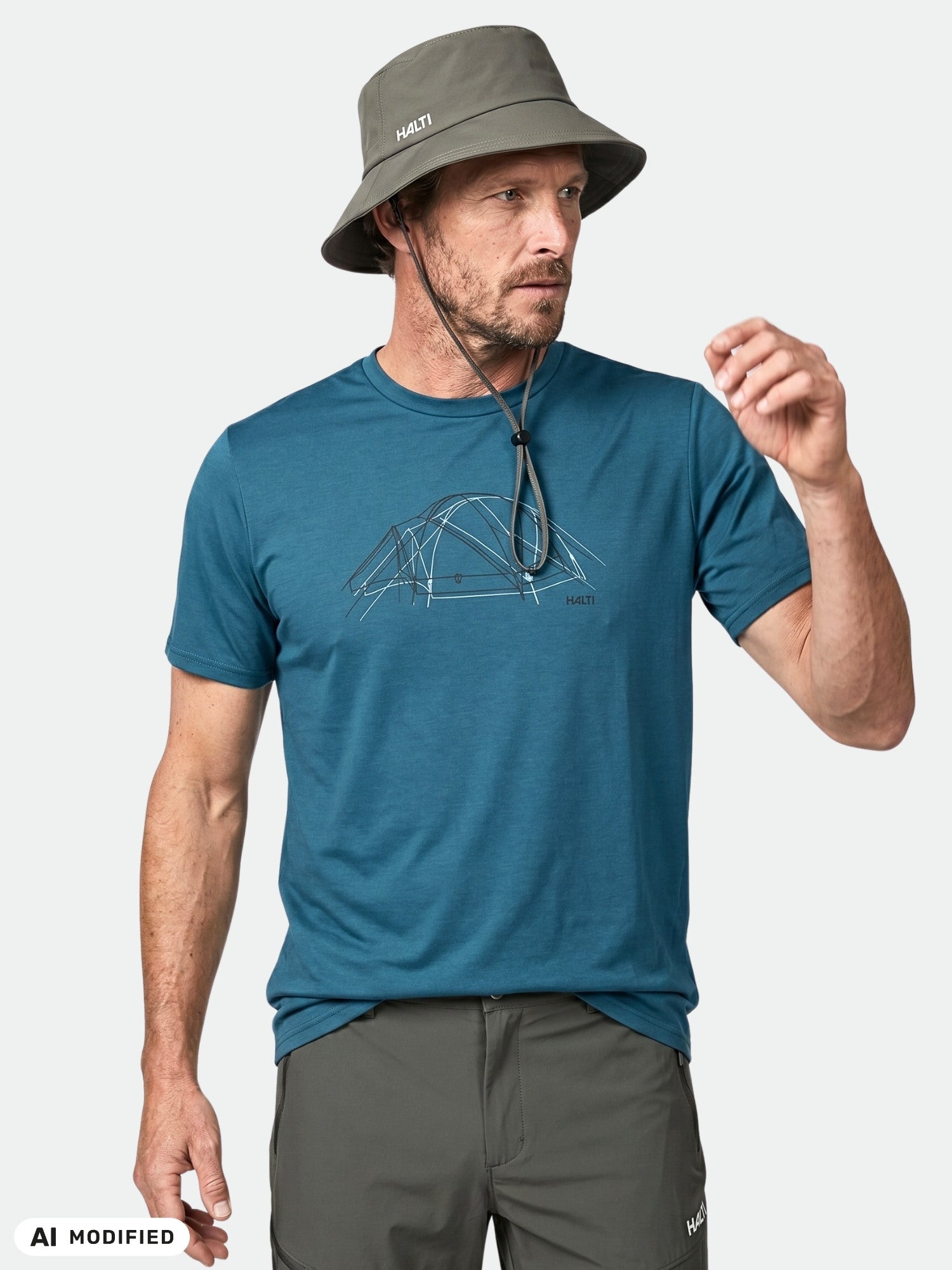 Man in Halti Kalteva T-shirt with tent graphic, gray bucket hat, and pants stands against a plain background.