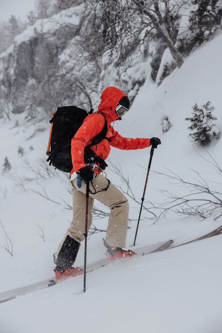 A woman in Halti Carvey II DX Ski Pants climbs uphill through snowy woods with skis, poles, and a red jacket.