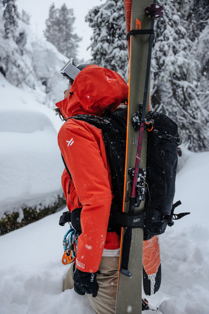 Woman in red jacket wears Halti Carvey II DX Ski Pants, standing in snowy forest with skis on her backpack.