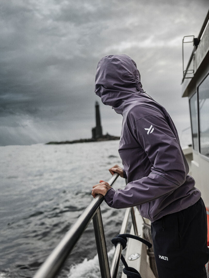 Wearing a Halti Staala DX Shell Jacket Women's, a person stands on a boat gazing at a lighthouse under cloudy skies.