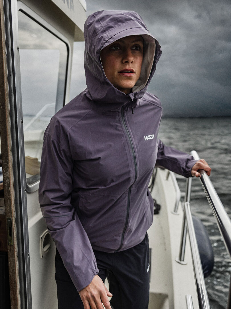A woman in a Halti Staala DX Shell Jacket stands on a boat, holding the rail with cloudy skies and water behind her.