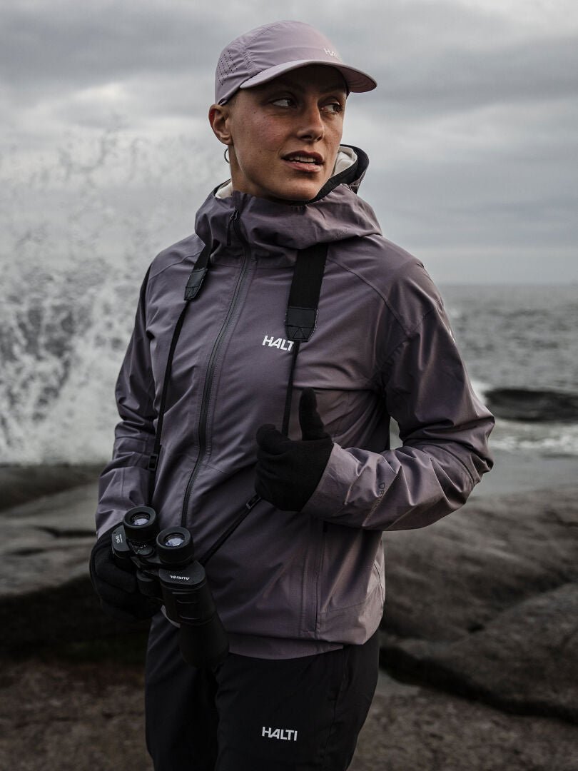 A woman in a purple Halti Staala DX Shell Jacket stands on a rocky shore with binoculars, waves in the background.