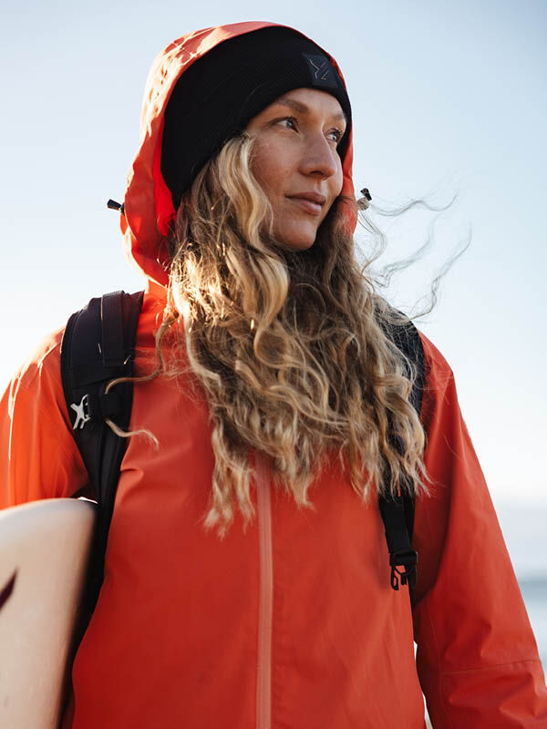 Woman in Halti Staala DX Shell Jacket and black beanie holds surfboard, wind blowing her hair outdoors.
