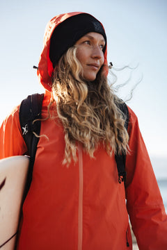 Wearing the Halti Staala DrymaxX Shell Jacket and a black beanie, she holds a surfboard and looks to the side outdoors.