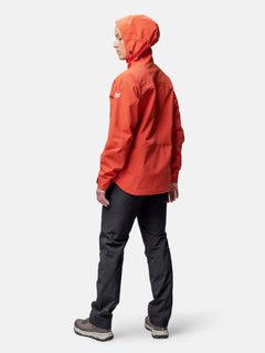 Woman wearing Halti Staala DrymaxX Shell Jacket in orange and dark pants stands facing left on a white background.