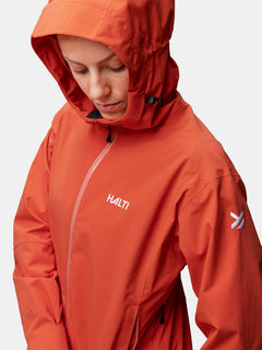 A person wears the Halti Staala DrymaxX Shell Jacket Womens in orange, looking down with a neutral expression.
