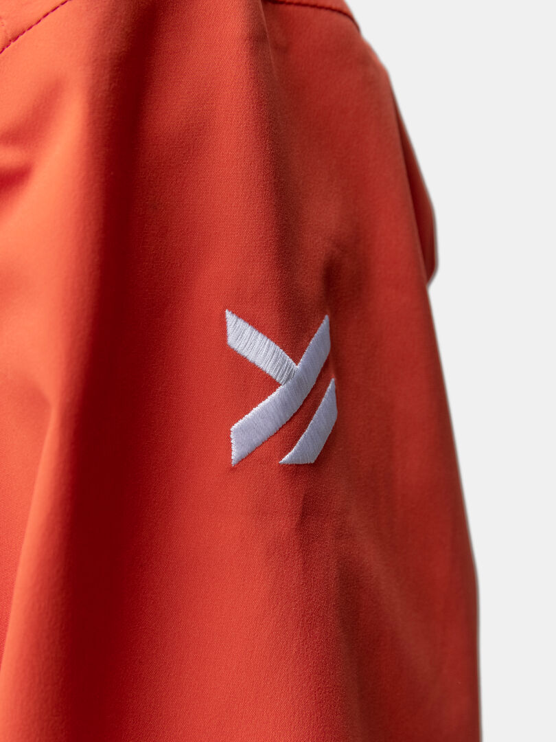 A white embroidered logo with two diagonal lines accents the sleeve of Haltis Staala DrymaxX Shell Jacket Womens in orange.