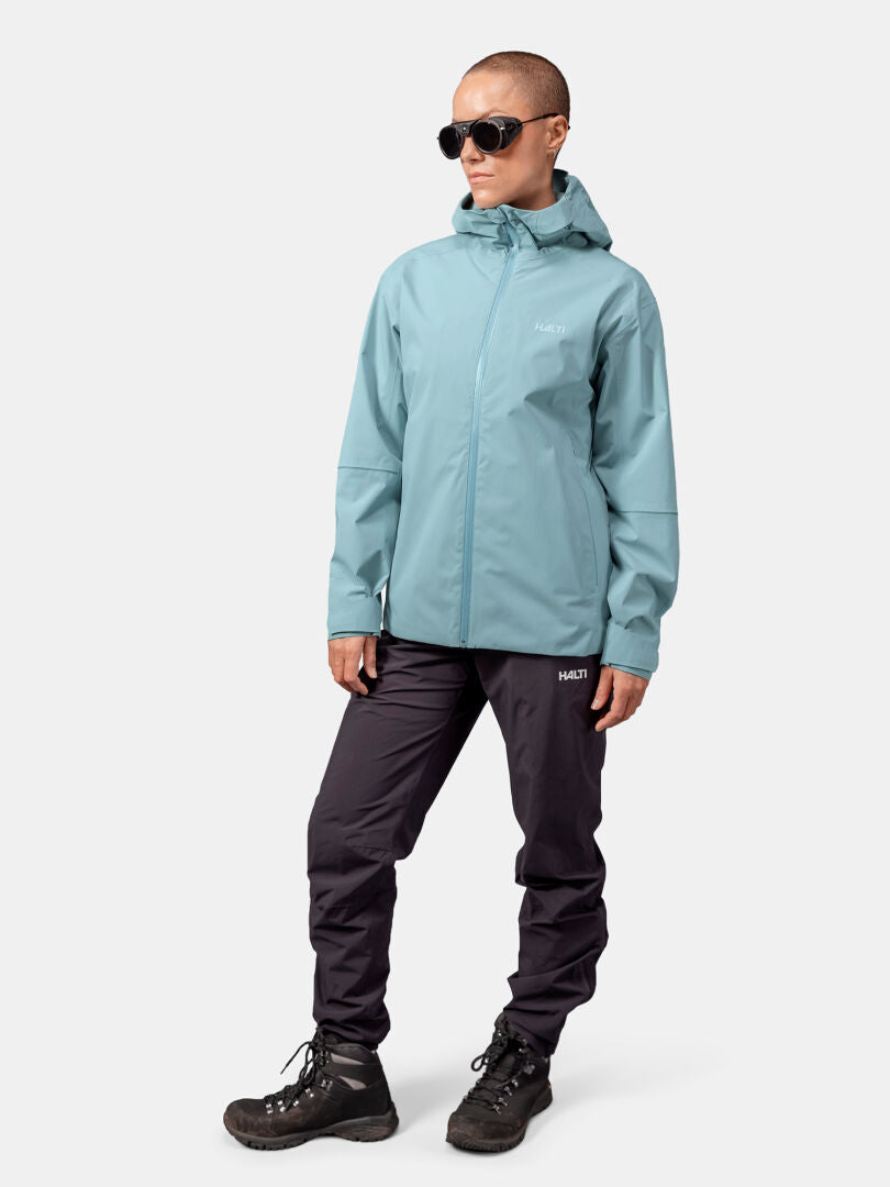 A person in a Halti Staala DX Shell Jacket Women's, sunglasses, black pants, and boots stands against a white background.