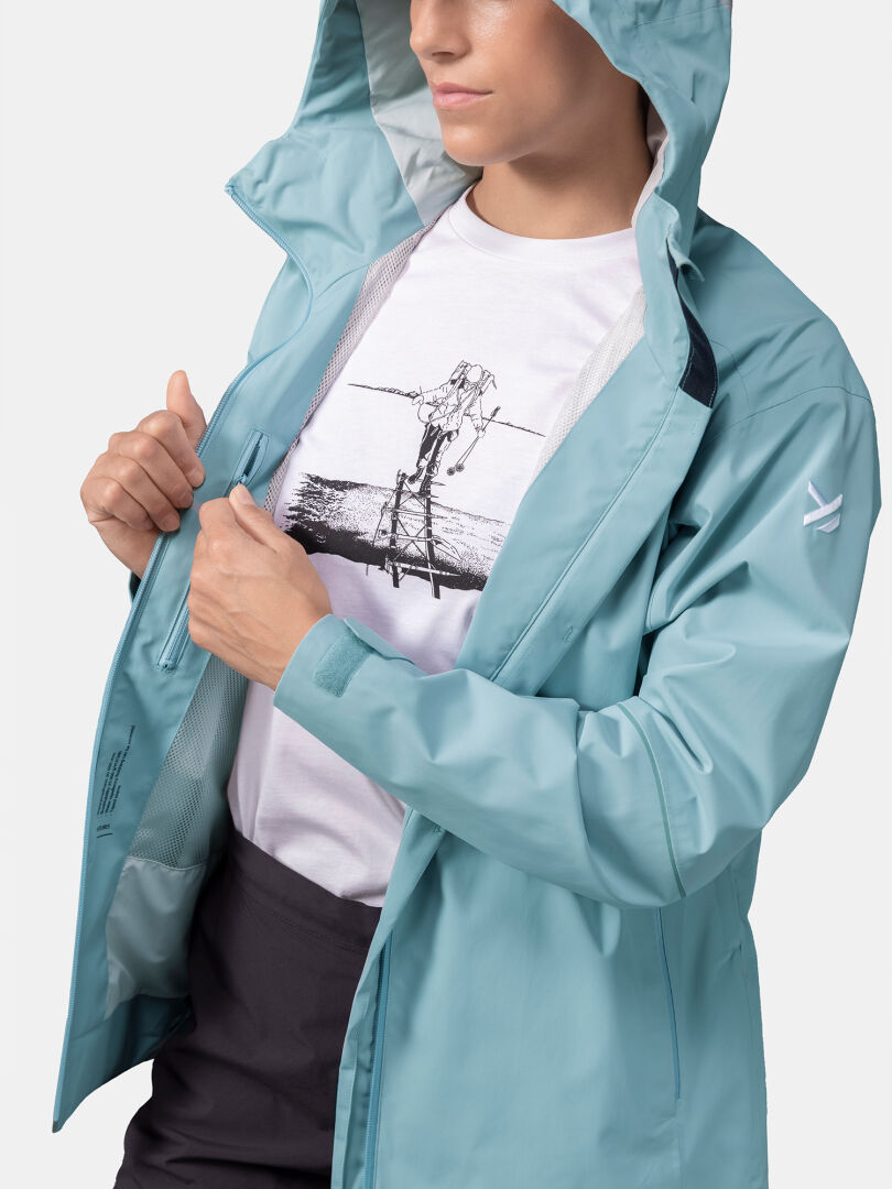 A woman in a light blue Halti Staala DX Shell Jacket unzips it to reveal a white tee with a black front graphic.