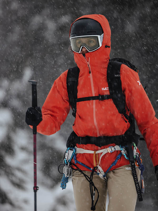 Woman in a Halti Staala DX Shell Jacket stands in falling snow, helmet and climbing gear visible.