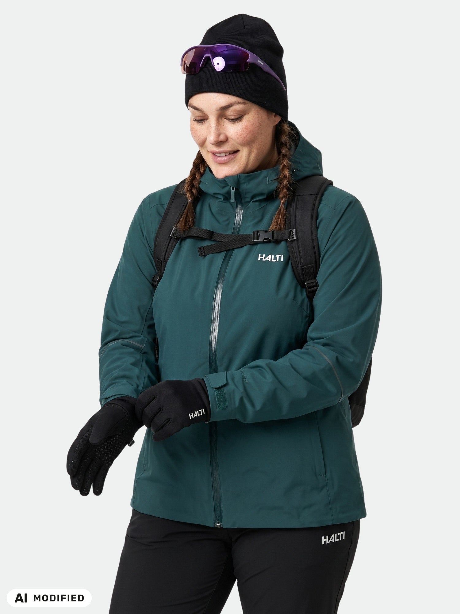 Woman in teal Halti Staala DX Shell Jacket Plus, black gloves, hat, sunglasses, adjusting glove, backpack straps visible.