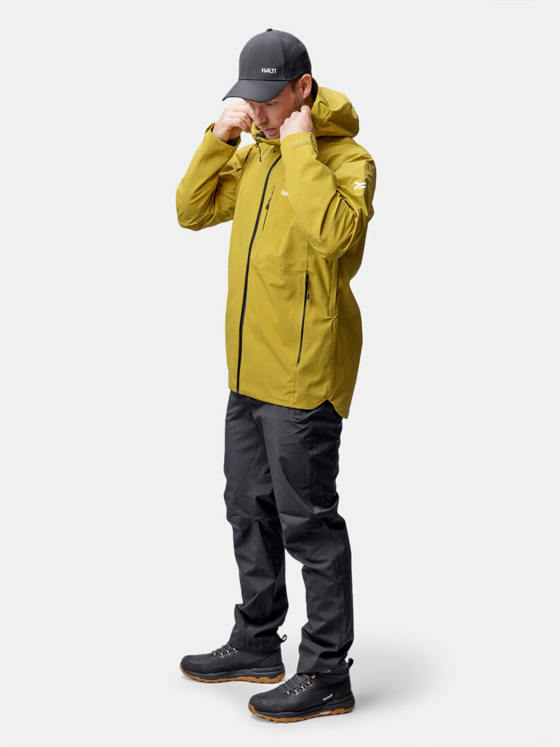 A man in a Halti Staala DX Shell Jacket adjusts his yellow hood while wearing a black cap, pants, and boots.