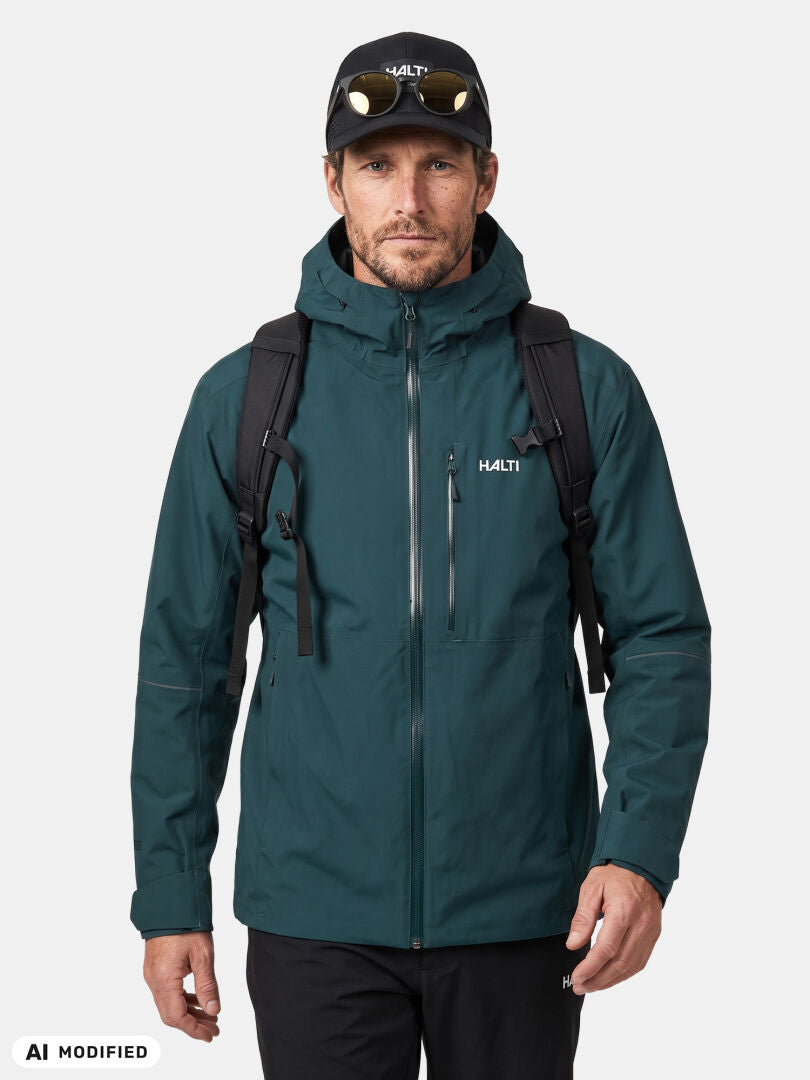 Man in Halti Staala DX Shell Jacket (teal), black backpack, and cap, standing against a plain white background.