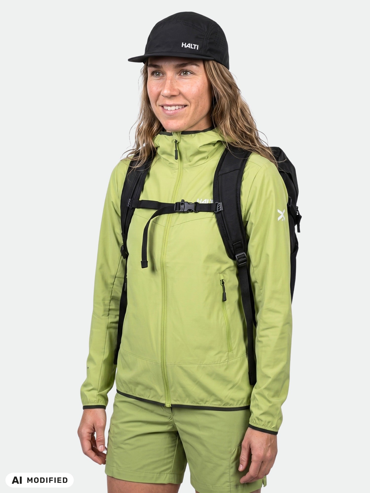 Woman in Halti Pallas Cool X-stretch green jacket, black cap, and backpack stands against a plain background.