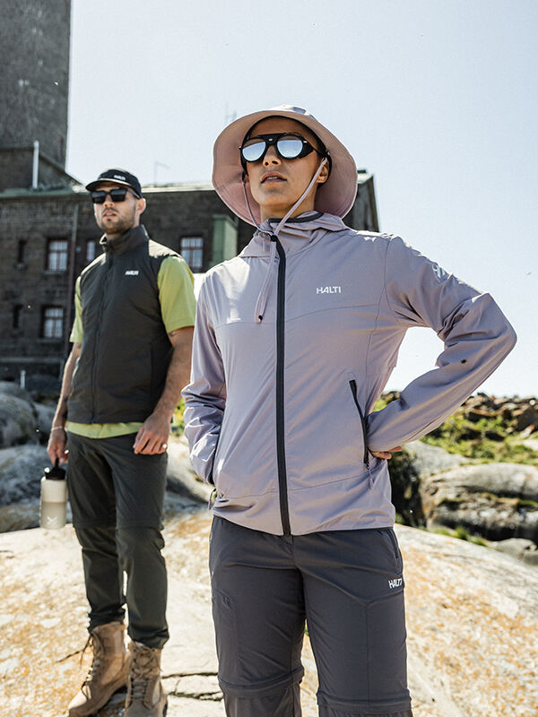 Two women in Halti Pallas Cool X-stretch Jackets and sunglasses stand on rocks by a stone building on a sunny day.
