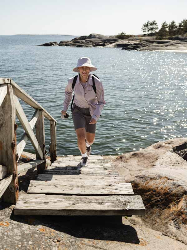Wearing Halti Pallas Cool Stretch Zip-off Pants, she climbs wooden steps from a rocky shore by the water with a backpack.