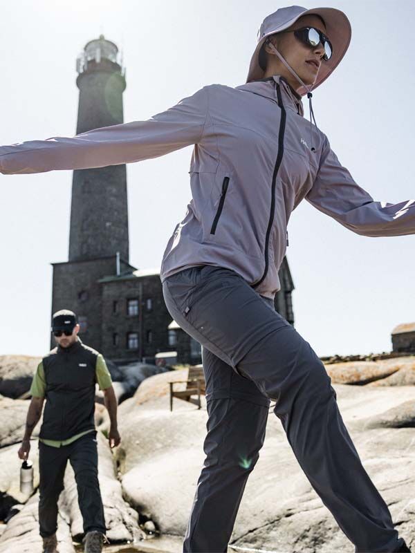 Two women in Halti Pallas Cool Stretch Zip-off Pants hike on rocks by a lighthouse and stone building under a clear sky.
