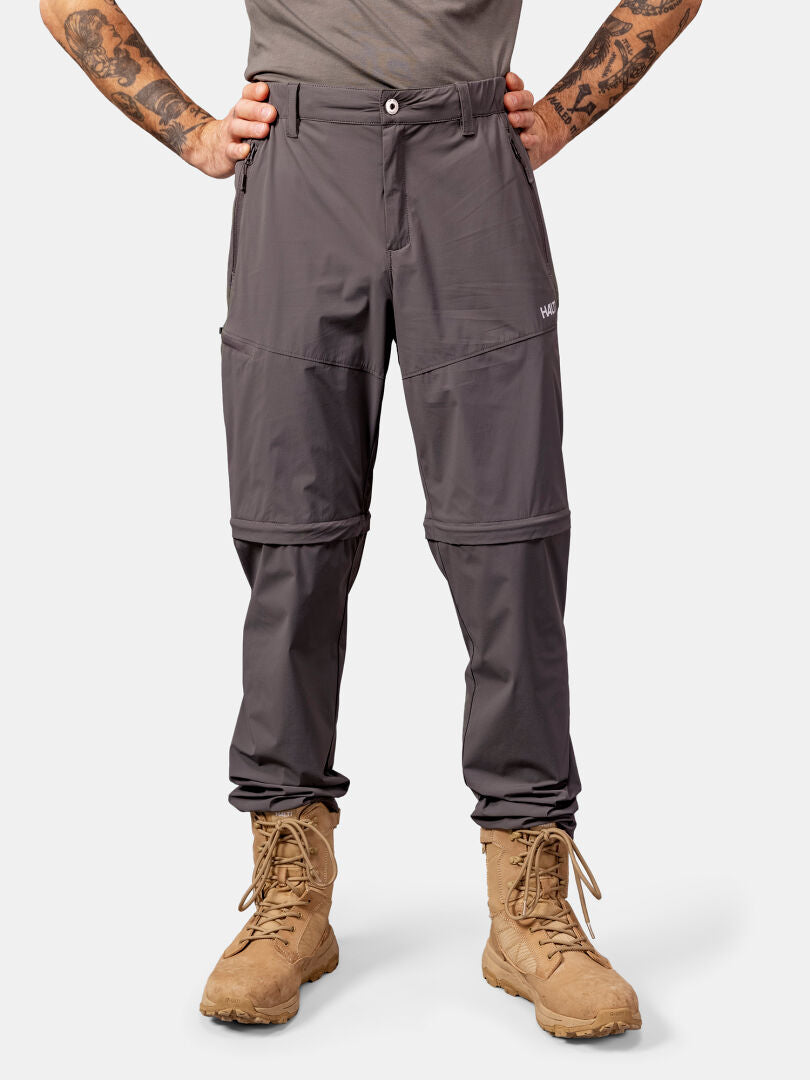 Man wearing Halti Pallas Cool Stretch Zip-off Pants in gray and tan combat boots, hands on hips, against a plain backdrop.