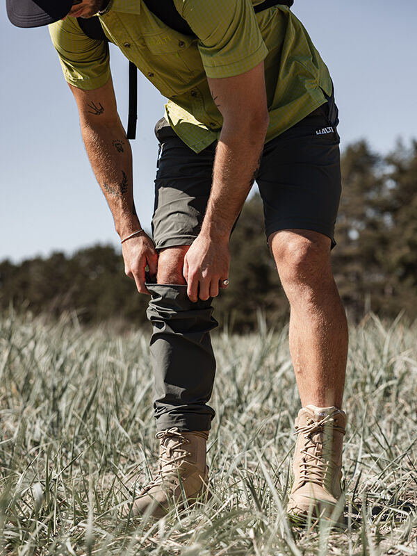 Wearing Halti Pallas Cool Stretch Zip-off Pants, a hiker pulls them over boots in a grassy outdoor area.
