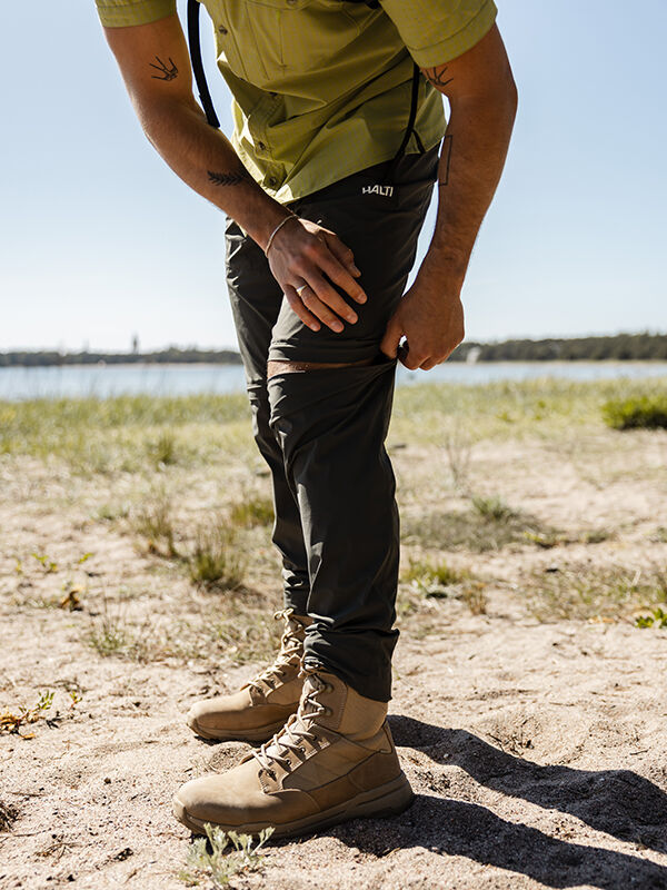 Wearing Halti's Pallas Cool Stretch Zip-off Pants, a man in outdoor gear stands by water on sandy ground.