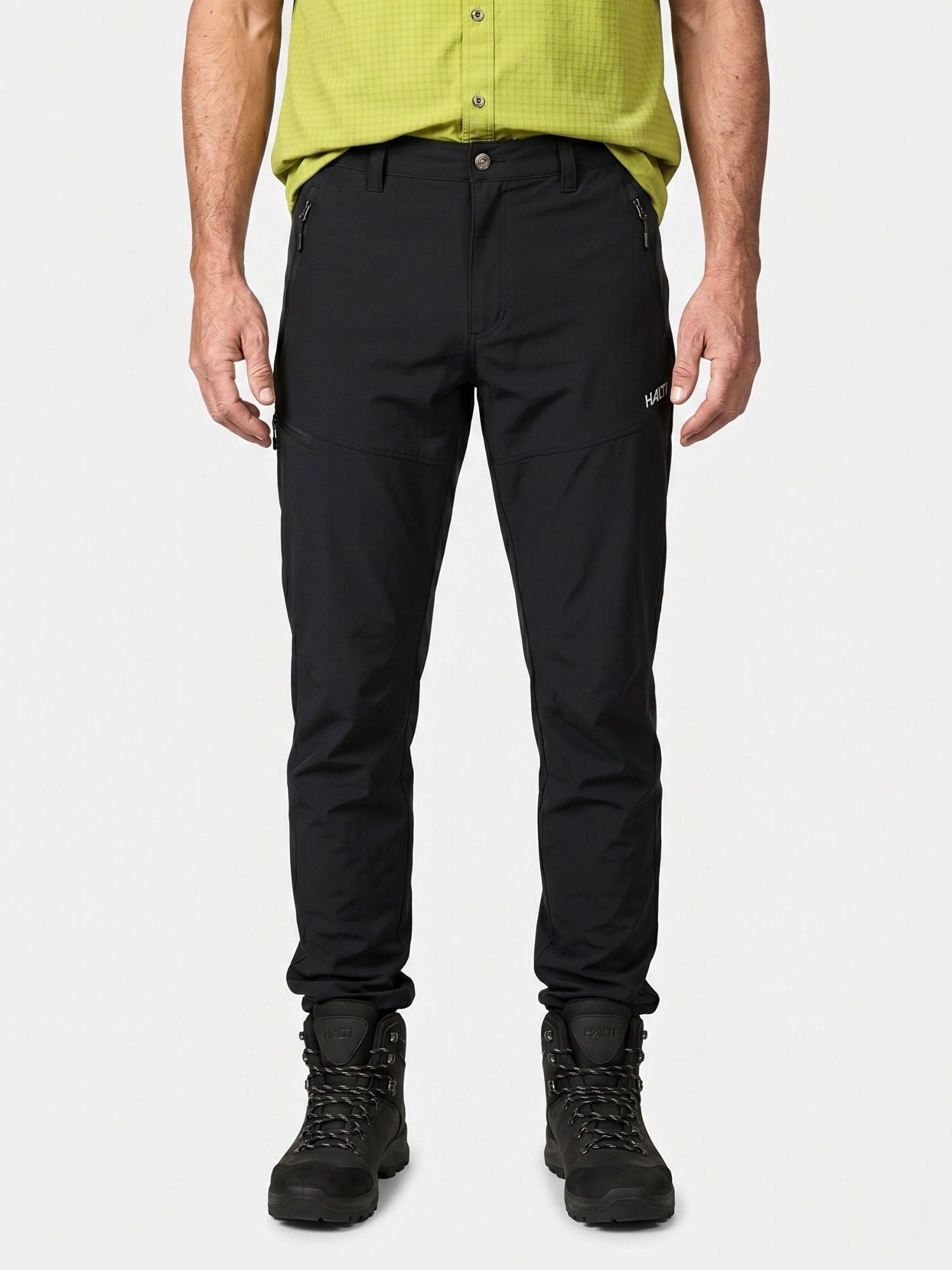 A person in Halti Pallas Cool X-stretch Pants, a light green shirt, and black hiking boots stands against a plain background.