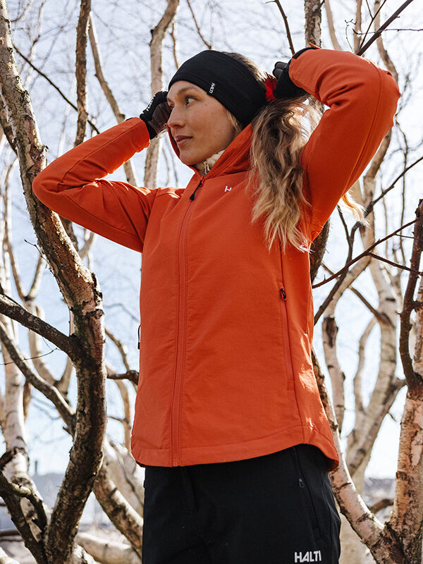 A woman in a Halti Finholma X-stretch Jacket adjusts her hair outdoors near leafless trees.