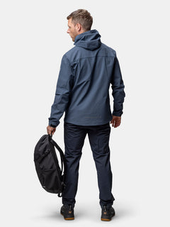 A person in a blue Halti Finholma X-stretch Jacket stands facing away, holding a black backpack in their left hand.