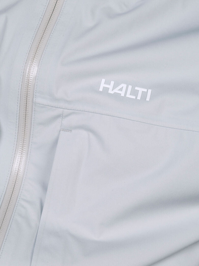 Close-up of the Halti Taival DX 3L Men’s Jacket in light gray, featuring a zipper and white chest logo.