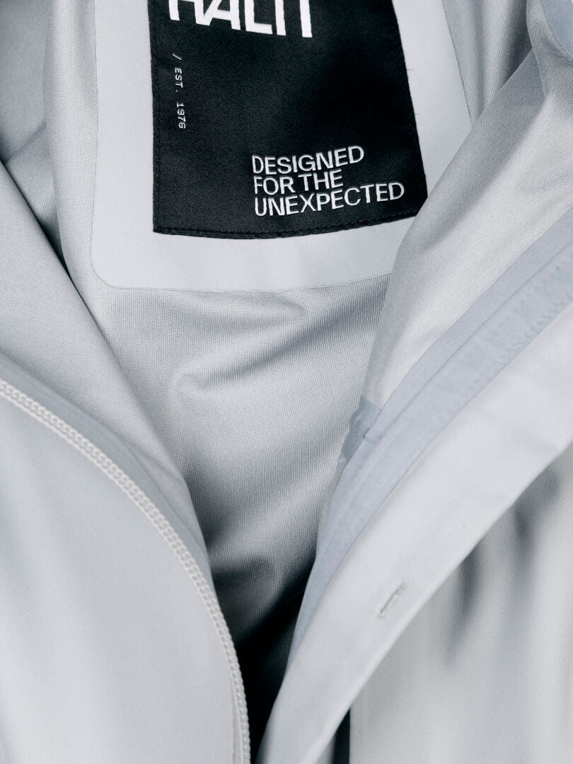 Close-up of the Halti Taival DX 3L men's jacket in light gray, featuring a black label: DESIGNED FOR THE UNEXPECTED.