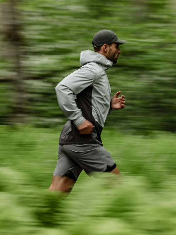 A man runs outdoors in the Halti Taival DX 3L Jacket Men's, with a green, blurred natural background.