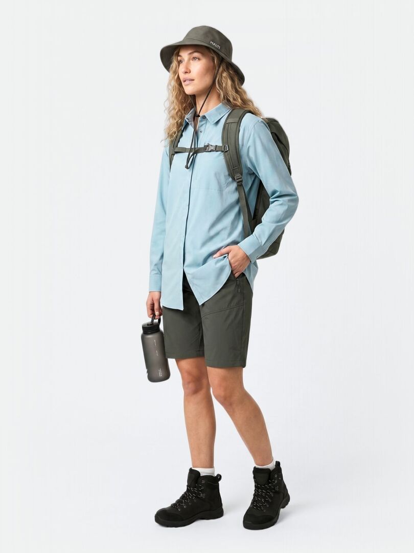 A woman in a Halti Samoy Longsleeve Trekking Shirt, hat, dark shorts, boots, backpack, holding a water bottle.