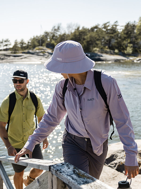 Two people wearing Halti Luovi Bucket Hats and quick-drying outdoor clothing walk by water, trees, and rocks in the sun.