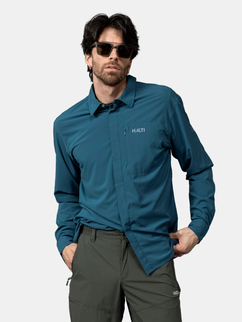 A man in sunglasses wears the Halti Samoy Longsleeve Trekking Shirt (UPF 90+, blue) and green pants, hand in pocket.